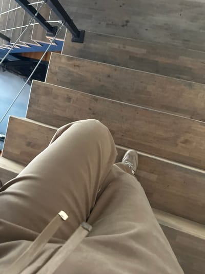 A first-person perspective photograph shows a person descending a wooden staircase. The person is wearing tan-colored pants and light-colored sneakers. The staircase has a black metal railing on the left side. Below the staircase, through the railing, a blue surface is visible. The location appears to be indoors, with potential evidence suggesting it could be in Marneuli, Georgia.