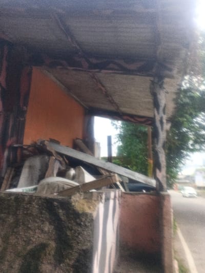 The scene is located in Dakshin Hichachhara, India, depicting a partially enclosed roadside structure. A dark, possibly corrugated, overhead canopy spans the upper frame, supported by a prominent dark-colored, textured pillar on the right. Below the canopy and to the left, a low concrete barrier or wall partially obstructs a collection of debris within the structure, consisting of wooden planks, assorted grey and white materials, and general refuse, positioned against an orange-brown interior wall. Through the structure's opening, bright, diffuse light from an overcast sky is visible, along with green tree foliage to the right. A paved asphalt road runs parallel to the structure on the right, where a light-colored vehicle is discernible in the blurred mid-distance, moving away from the viewer. No individuals are present in the frame.