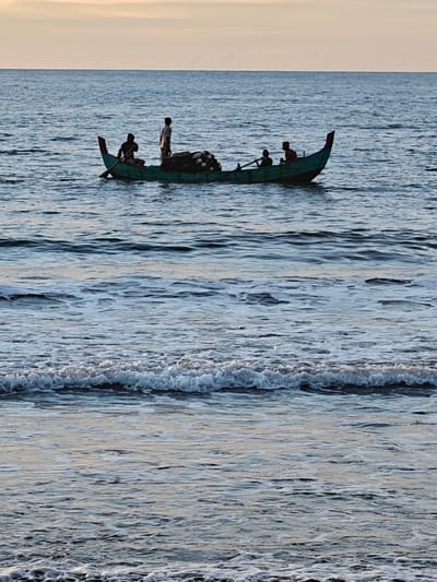 The image captures a serene scene at sea, likely near Uleekareung, Indonesia, given the context. A narrow, teal-colored boat is navigated by four figures. One individual, standing, appears to be observing the horizon, while three others are seated. One of the seated figures is actively rowing with a single oar. The boat carries a substantial load of fishing nets, piled high and covered. The water is a deep blue, with gentle waves creating a textured surface. In the foreground, smaller waves with foamy crests approach the viewer. The sky above is a soft gradient of pale yellow and blue, suggesting either sunrise or sunset. The overall atmosphere is one of calm and industry.