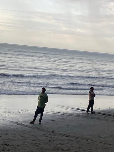 The image shows a beach scene with two individuals standing on the wet sand near the water's edge. The ocean has moderate waves and the sky is overcast with clouds. One person, dressed in a green long-sleeved shirt and dark pants, is closer to the viewer and facing towards the water with their body angled away. Their left leg is extended forward, and they appear to be leaning. The second person, wearing a light-colored short-sleeved shirt and dark pants, is further down the beach and is facing away from the viewer, with their arms crossed. The sand shows some tire tracks or similar markings, possibly from a vehicle. The overall lighting suggests it could be early morning or late afternoon. This scene is located on the beach in Bateetimoh, Indonesia.