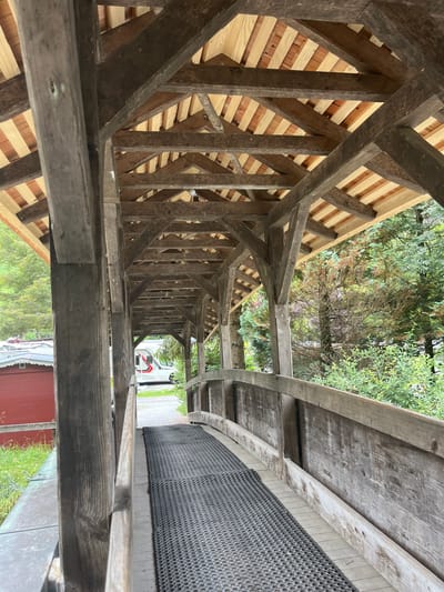 The image shows a covered wooden bridge in Wengen, Switzerland. The bridge has a wooden roof supported by a timber frame. The walkway is covered with a black rubber mat. To the left of the bridge, a red building with a white roof is visible, and beyond that, lush green foliage. In the background, a white camper van with red and black accents is parked on a paved area. The environment appears to be a rural or mountainous setting with dense trees and vegetation.