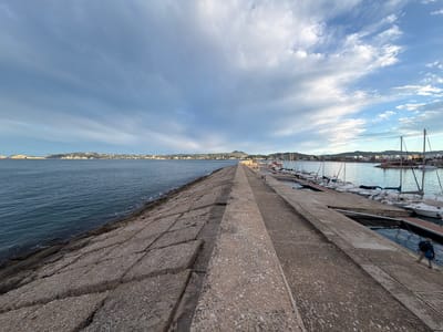 This is an eye-level, wide shot of the breakwater at Xàbia/Jávea, Spain. The breakwater is made of large, rough, gray stones on the left and a smoother concrete surface on the right. In the distance, there's a stretch of land with buildings, and a prominent hill visible. The water is relatively calm. Several white sailboats are moored on the right side of the breakwater. A man wearing a backpack walks along the concrete path, away from the viewer. The sky is filled with clouds, ranging from dark and gray to light and white. The overall light suggests it might be early morning or late afternoon.