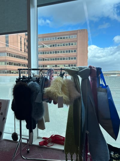 The image captures an indoor perspective looking out through a large window onto an urban setting in Garston, United Kingdom. In the immediate foreground, a chrome-plated mobile clothing rack is positioned, displaying a collection of winter accessories. These include several knitted hats, some featuring prominent fur pom-poms in dark brown and light grey, alongside other knitted items. Multiple long, knitted scarves hang from the rack, presenting in colors such as light green, light blue, and pink, with some exhibiting fringed edges. A fluffy yellow accessory, possibly a collar or scarf, is also visible, with a retail tag attached. To the far right, a blue and grey reusable shopping bag is suspended from the rack. On the white windowsill beneath the window, a red pouch or small bag, branded with "MOSCHINO," lies horizontally. The floor in the foreground is covered with a reddish-purple patterned carpet.
Beyond the window, under a sky with patches of blue and white cumulus clouds, two large, multi-story urban buildings are visible. Both structures are constructed from a reddish-brown material, likely brick or concrete, and feature numerous rectangular windows arranged in consistent rows. One building stands prominently across the view, while a section of a similar building is visible to its left. A light grey roller blind is partially lowered at the top left of the window frame. No individuals are present in the scene.