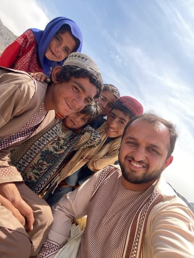 A low-angle, outdoor medium shot shows six individuals, including one adult male in the foreground, possibly taking a selfie in the city of Ashoqah, Afghanistan. The man in the foreground is smiling and wearing a long-sleeved, patterned tan shirt. Behind him are five children of varying ages; one child is wearing a light-colored embroidered cap, another a red embroidered cap. One child is positioned behind the man in the foreground. A girl wearing a red shirt and blue headscarf is to the left of the man. The background suggests a mountain landscape under a bright, partly cloudy sky. The subjects appear to be smiling and engaged with the camera.