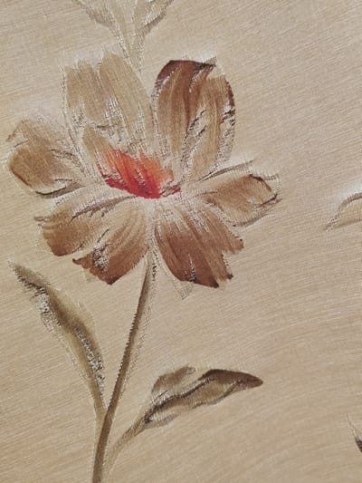 This is a close-up, high-angle, full-frame photograph of a textured fabric with a painted floral design. The background fabric is a light beige with a subtle weave that creates a faint horizontal and diagonal texture. The primary subject is a single, stylized flower rendered in shades of brown and a vibrant red-orange at its center. The flower has five prominent petals with visible brushstrokes, appearing somewhat abstract and impressionistic. The petals are outlined with darker brown strokes, and some have highlights of lighter beige. A thin, brown stem extends downwards from the flower, with two elongated leaves painted in similar shades of brown and beige. The overall impression is of a decorative textile, possibly upholstery or drapery. The location in the city of Gardene, Latvia, is not determinable from the image.