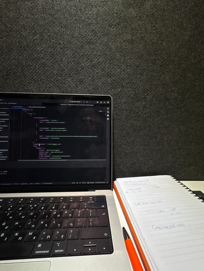 The image depicts an indoor workspace located in Nes Ziona, Israel. A silver-grey Apple MacBook laptop is open on a white desk surface, positioned towards the left of the frame. Its screen displays a code editor, likely Visual Studio Code, showing a JSON configuration file named "testConfig.json" with code fragments including "create BZON user", "headers", "application/json", and "ANDROID_ID". The laptop's keyboard features both English QWERTY and Hebrew characters. To the right of the laptop, a white spiral-bound notebook with an orange spine is open on the desk, displaying handwritten notes in English. Visible text includes "Get Mode Supply Request", "Response", "Get full line info", "WEB", "PDF", and "IP 192.168". An orange pen with a black tip rests on the open notebook. The background consists of a dark, textured wall or partition. No individuals are present.