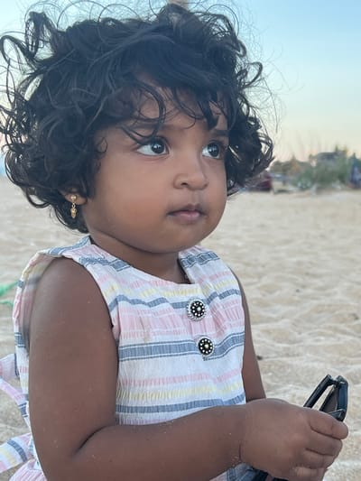 A child is sitting on a beach. The child has dark, curly hair and brown skin. The child is wearing a light-colored, striped dress with decorative buttons. The child is holding a black object, possibly sunglasses. The background shows a sandy beach and a blurry view of buildings. The location is Kalmunai, Sri Lanka.