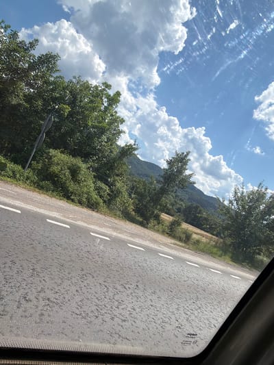 The image captures a scenic view from inside a vehicle traveling on a road, likely near Mikre, Bulgaria. The asphalt road occupies the foreground, with white dashed lines marking its lanes. Beyond the road, lush green trees and vegetation line the slope, leading up to a mountainous landscape in the background. The sky is a vibrant blue, dotted with fluffy white clouds and streaks of sunlight reflecting off the vehicle's windshield. A road sign is visible on the left side of the image. The overall impression is one of a peaceful, natural environment observed during a journey.