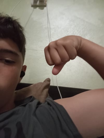 The media file shows a close-up, selfie-style shot of a young person. The subject is looking towards the camera with a slightly apprehensive or uncertain expression. They have dark hair and are wearing a dark t-shirt and a black earbud. Their right arm is raised, with their hand forming a thumbs-down gesture. The background appears to be an indoor setting with a textured, light-colored wall. A white electrical outlet with two sockets is visible on the wall near the top of the frame, and a thin white cord hangs down from it. The lighting suggests it is nighttime or indoors with artificial light. There is no visible text in the image. The overall mood is casual and somewhat informal. The subject's pose and expression suggest a negative reaction or disapproval.