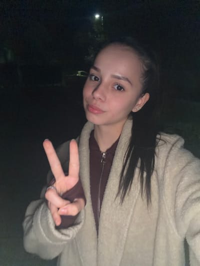 A young woman with dark hair pulled back into a ponytail is facing the camera, making a peace sign with her right hand. She is wearing a maroon sweater with a silver zipper and a light beige, possibly fleece, jacket. The background is very dark, suggesting nighttime or a dimly lit area. There is a hint of greenery and a faint light source in the distance. The location in the city of Bov, Bulgaria is not determinable from the visual elements of the image.