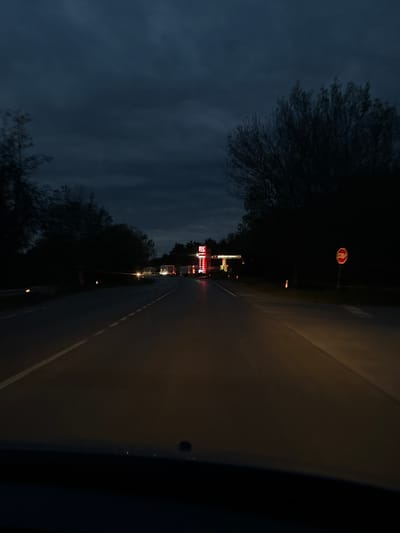 The image presents a nighttime or late dusk scene from the perspective of a vehicle traveling on a road in Momin Sbor, Bulgaria. The sky is dark and overcast, with silhouettes of trees visible against it on the left side of the road.

The road ahead is a dark asphalt surface, with a faint white line marking its right edge. On the left, further down the road, several commercial buildings are brightly lit with various neon signs. A prominent red octagonal stop sign stands out against the dark background, positioned above the buildings.

Among the illuminated structures, a recognizable red and white sign for "BURGER KING" is clearly visible, with its distinct branding and lettering. Below it, another red sign, though less clear, appears to indicate a "DRIVE THRU" service. Other smaller lights and light sources, possibly from streetlights or other vehicles, dot the distant scene. The partial view of the vehicle's interior (dashboard or windshield frame) in the foreground suggests the image was captured by a driver or passenger. The overall atmosphere is one of a typical commercial roadside environment after dark.
