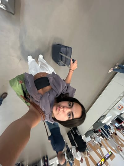 The image shows a young woman with long dark hair taking a selfie while pulling a dark blue suitcase with her right hand. She is wearing a light gray sweatshirt, white pants, and a black sports bra. She is standing on a white tiled floor and has a green bag hanging on her left shoulder. The woman is looking directly at the camera. In the background, a long line of people are waiting in a queue at an airport check-in. The coordinates (latitude: 28.047, longitude: -16.58) suggest that this location is in Granadilla de Abona, a town in the south of Tenerife, Spain. The environment is an airport, with check-in counters visible in the distance. The overall scene is one of travel and anticipation.