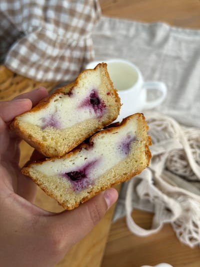A close-up shot shows a hand holding a sliced baked good. The baked good is golden brown on the outside and has a creamy white filling studded with dark purple berries, likely blueberries. The texture appears moist and dense. In the background, out of focus, is a white mug and a light-colored fabric with a checkered pattern, as well as a loosely woven white fabric. The overall setting suggests a casual indoor environment, possibly a kitchen or dining area, and the lighting implies it could be daytime. No text is visible in the image.