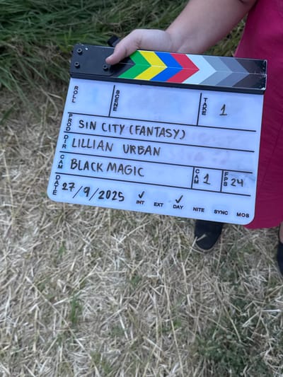 An individual holds a film clapperboard in an outdoor, grassy environment in El Paraíso, Mexico. The clapperboard displays the following information: "PROD / DR: SIN CITY (FANTASY)," "Director: LILLIAN URBAN," "Camera: BLACK MAGIC," "TAKE: 1," "CAM: 1," "FPS: 24," and "DATE: 27/9/2025." Checkmarks indicate "INT" (Interior) and "DAY," with "EXT" (Exterior) unchecked, contradicting the visible uncultivated field of dry, tan, and green grass that comprises the background. The person holding the clapperboard is partially visible, wearing a red garment and dark footwear. The hand gripping the clapperboard is in the upper center, and a lower leg and foot are visible in the lower right portion of the frame.