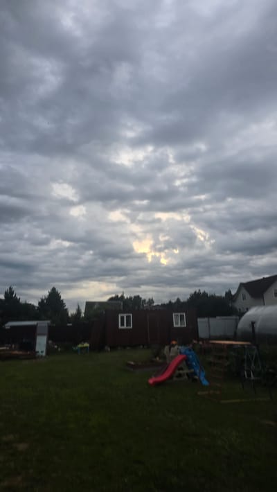 The image depicts a backyard scene in Strokino, Russia, under a heavily overcast sky with patches of sunlight breaking through the clouds. In the foreground, a well-maintained green lawn occupies most of the space. Scattered across the lawn are various children's play structures, including a red and blue slide, a sandbox, and a jungle gym. A dark brown shed or outbuilding with two white-framed windows and a single door is visible in the middle ground, positioned behind the play area. To the left of the shed, a long, low structure with a white roof and dark siding is partially visible. Beyond the shed and play area, a dense line of trees forms a backdrop, with the roofline of a residential house peeking out from behind them on the far right. A greenhouse-like structure is also present on the far right side of the image. The overall impression is one of a suburban or rural residential area, likely a dacha or country home, on a cloudy day.
