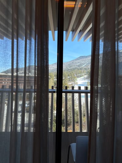 The image shows a view from indoors through a window with semi-transparent curtains and wooden slats. The view is partially obscured by the curtains and slats. Outside, there is a scene of mountains, trees, and possibly a body of water. There is also a balcony or deck visible with a railing and a white chair. It is not possible to determine the location as Озёрное, Russia, without further information.