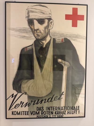 This media file is a photograph of a framed poster. The poster itself is a vintage graphic artwork depicting a man with a bandaged head, leaning forward with his eyes closed. The style of the illustration is rough, with bold lines and a limited color palette dominated by black, white, and a touch of red.
On the left side of the poster, a red cross symbol is prominently displayed against a white background. To the right of the illustration, vertical text is printed in a dark color. The largest text, written in a flowing script, reads "Verwundet," which is German for "Wounded." Below this, in a more formal, sans-serif font, is the text "DAS INTERNATIONALE KOMITEE VOM ROTEN KREUZ HILFT!", translating to "The International Committee of the Red Cross Helps!". Further down, there is smaller text indicating "POSTCHEK I. 777 GENF" and "J.C.MÜLLER ZÜRICH 8".
The overall mood of the poster is somber and serious, likely intended as a public service announcement or appeal for support for the Red Cross and its efforts to aid the wounded. The poster is framed in a simple black frame and appears to be hanging on a white wall, suggesting it is displayed in an exhibition or a similar public space. The reflection on the poster's glass indicates it is well-lit, possibly with artificial lighting. The poster's content and its displayed context point towards a historical or informational setting.