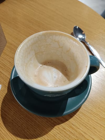 A close-up, high-angle shot shows a nearly empty, white-rimmed teal coffee cup with latte art residue on a matching teal saucer. The cup sits on a light brown wooden table. A silver spoon rests on the table to the right of the cup. The background is blurred. The image is located in Buzău, Romania.