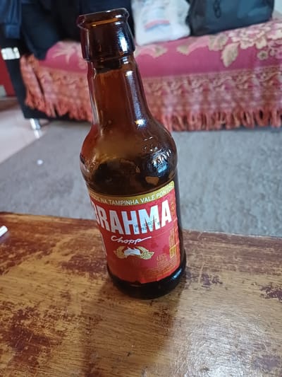 The image shows a close-up of a brown glass beer bottle lying on its side on a wooden surface. The bottle has a red label with white text. The visible text on the label includes "BRAHMA," "Chopp," "4.8%," and "CERVEJA." There is also some smaller text, including what appears to be a date code and information about containing gluten. The bottle's cap is visible, and the label has a design with what looks like a hop icon. The wooden surface is textured and appears worn. In the background, out of focus, is a patterned fabric, possibly a blanket or rug, with pink and floral designs, and a textured gray surface. The lighting suggests an indoor setting, possibly during the day. No people are visible in the image. The scene appears to be a casual setting, with the beer bottle as the central object. The context of Itabira, Brazil, suggests this could be a common sight in a home or bar in the region.