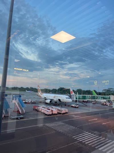 The image depicts an airport scene viewed through a window, likely at Ramunia Dua, Indonesia. There are multiple aircraft parked at gates with jet bridges connected, including a Super Air Jet airplane. Ground crew equipment such as luggage carts and passenger stairs are visible next to the planes. The sky is cloudy, reflecting in the window, and the terminal building reflects "Plaza Premium" in reverse. The ground is marked with runway markings and pedestrian crosswalks.