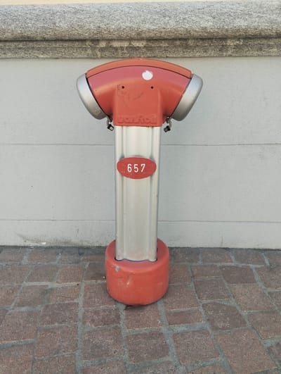 The image displays a modern, T-shaped fire hydrant standing against a light grey wall and cobblestone ground. The hydrant is predominantly red and silver. Its rounded top section is red with silver caps on either side, resembling a head with ears. The word "VonRoll" is embossed or printed in white letters on this red top section. Below the red top, the main body of the hydrant is silver. A red oval plate, prominently featuring the number "657" in white, is attached to the silver column. The base of the hydrant is also red and cylindrical.
The hydrant appears to be in an urban setting, possibly a sidewalk or public square in Bellinzona, Switzerland, given the provided context. The wall behind it is textured and light grey, with subtle horizontal lines. Above the wall, a rough-hewn, grey stone ledge or trim runs horizontally across the upper portion of the frame. The ground beneath the hydrant is paved with reddish-brown, somewhat irregular cobblestones or pavers, separated by grey mortar lines. The lighting is bright and even, indicating it is daytime. There are no people or discernible activity in the scene. The hydrant itself shows some minor wear and tear, with a few scuffs and scratches visible on its surface.