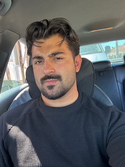 A man with dark hair and a beard is sitting in a car. He is wearing a dark-colored sweater. The car is parked, and the view through the window shows a building and possibly some scaffolding. The image's angle suggests the photo was taken as a selfie. The specific location in Dolni Tsibar, Bulgaria, cannot be determined from the visual data.