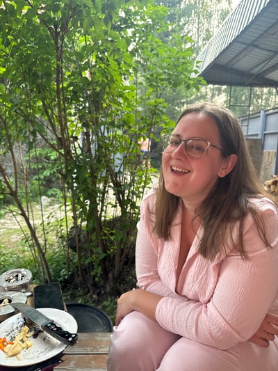 A medium shot shows a woman with shoulder-length brown hair and glasses smiling broadly. She is wearing a light pink textured outfit. She is seated at a wooden table outdoors. In the foreground, on the table, are two ashtrays with cigarette butts, a smartphone, a plate with food scraps and a knife. Behind her, there is lush green foliage and a corrugated metal roof structure. In the background, to the left, a partially visible person in swim trunks can be seen among the trees. The scene appears to be in the city of Ust-Mana, Russia, possibly at a dacha or outdoor recreational area.