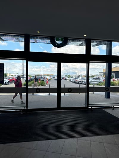 The image captures an indoor view looking out through a large set of glass doors. The perspective suggests the photographer is standing inside a building, facing an exit.

**Subjects:**
One person, wearing a red top and light-colored pants, is prominently visible walking towards the right side of the frame, seemingly exiting the building through the glass doors. Another person in a light top and dark pants is visible further outside in the parking lot. The outdoor scene features a large parking lot filled with numerous parked cars of various colors, including white, silver, and red. Beyond the parking lot, modern buildings are discernible under a bright sky. Inside, a dark ceiling with a small light fixture is visible, and the floor features a large black ribbed mat leading up to the doors, transitioning to light grey tiles.

**Activity or event taking place:**
The primary activity shown is a person exiting the building. Other individuals can be seen walking or standing within the parking lot, suggesting typical daily activity at a commercial center.

**Notable details:**
The scene is set during the daytime, indicated by the bright sky and clear visibility outdoors. The weather appears fair, with white clouds scattered across a blue sky. A green "EXIT" sign, adorned with a dark green wreath, is mounted above the glass doors, likely indicating a holiday or seasonal decoration. The location is clearly identified by the text "ST GERMAIN CENTRAL" printed vertically on the dark frames of the glass doors. This aligns with the provided context of Clyde North, Australia, where St Germain Central is a known shopping and lifestyle precinct.

**Visible text:**
The text "EXIT" is clearly visible on the green emergency sign. The words "ST GERMAIN CENTRAL" are repeatedly printed vertically on the glass door frames.