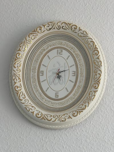 This is a medium shot, eye-level photograph of a decorative wall clock against a textured white wall. The clock has an ornate, oval design with a white frame featuring gold, scroll-like embellishments. The clock face is white and appears to be made of ceramic or a similar material, with Arabic calligraphy encircling the numbers. The numbers themselves are represented by small diamond-like studs and also by stylized Arabic numerals in a square format, with a prominent "9" visible at the bottom left. The clock hands are black and red, and it appears to be showing a time of approximately 10:10. In the center of the clock face, there is a raised, white relief of Arabic script. The lighting suggests it is daytime, likely indoors, given the consistent light on the wall and clock. The setting is a wall in what appears to be a residential or public space in Dorsten, Germany. There are no people visible, and no specific weather or emotional cues are present. The prominent text is in Arabic calligraphy around the clock face and in the central relief.