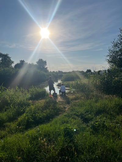 The image shows two figures fishing in a narrow body of water, possibly a river or canal, in a rural or semi-rural setting. The sun is bright and positioned in the upper left quadrant of the image, creating lens flare and casting strong light. The foreground is dominated by tall, green grass and reeds, suggesting a natural environment. In the mid-ground, a person wearing a dark t-shirt and shorts stands casting a fishing rod, with a bucket at their feet. To their right, a second person, wearing a light-colored t-shirt and shorts, is seated, also fishing. The water is calm and reflects the sky and surrounding vegetation. The background consists of dense trees and shrubs on either side of the water, with a few structures, including what appear to be outbuildings or temporary structures, visible in the distance on the right. The sky is blue with wispy clouds. The scene depicts a peaceful outdoor activity, likely during daytime. The location in Яново-Грушевский, Russia, is inferred from the provided context, but the visual elements themselves are generic to many temperate rural landscapes.