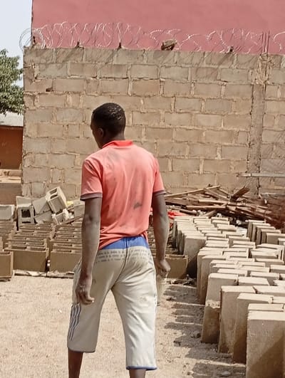 A man in a red t-shirt and khaki shorts stands with his back to the viewer. He is surrounded by rows of rectangular, light-colored bricks or blocks on the ground. Behind him is a brick wall topped with coiled barbed wire, and a red building is partially visible above the wall. A small pile of lumber rests near the bricks. The ground is covered in light-colored gravel.
