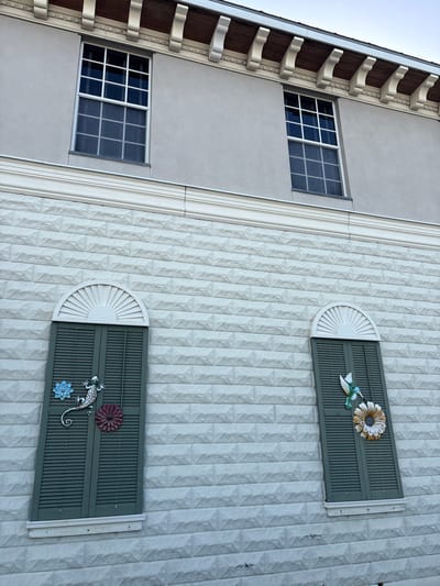 The image shows a partial view of a building's facade in Punta Gorda, United States of America. The building has two upper windows with white grids and dark panes, set against a light gray stucco wall. Below these windows, a decorative white molding separates the upper wall from the lower section. The lower section of the building is covered in white brickwork, with each brick having a beveled edge, creating a textured pattern.
There are two sets of dark green louvered shutters, each with a semicircular fanlight above. Both shutters are decorated with colorful metal ornaments. The left shutter features a silver lizard in the center, with a blue flower to its left and a deep red flower to its right. The right shutter has a green and blue hummingbird in flight on the left side, and a large, light-colored flower with a dark center below it. The building appears to be well-maintained, and the sky above is a pale blue.