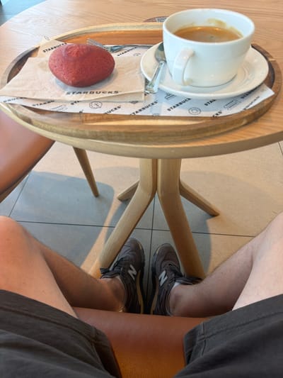 The image shows a person sitting at a small, round wooden table, likely at a café. On the table, there is a white coffee cup and saucer, a spoon, a small red pastry (possibly a brioche), and Starbucks-branded napkins. The person is wearing grey shorts and brown New Balance running shoes. The background consists of tiled flooring. The location is likely a Starbucks, but the specific location in Weil am Rhein, Germany cannot be confirmed from the image alone.