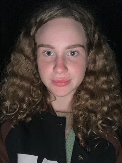A close-up, rotated selfie of a young woman with curly blonde hair and light blue eyes. She has fair skin and is smiling slightly. She is wearing a black jacket with a white emblem on the right side, layered over a beige sweater and a green shirt or undershirt. The background is dark and indistinct. No information indicates that this photograph was taken in Krasnyi Jar, Russia.
