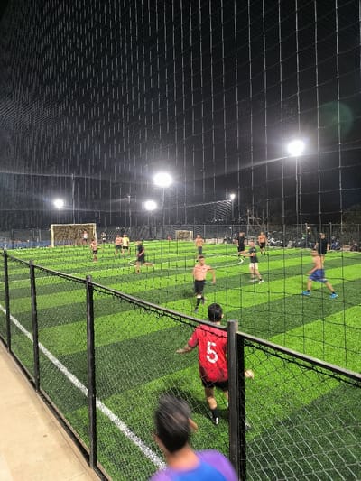 The image captures an evening or night-time football (soccer) game played on an artificial turf pitch, illuminated by bright floodlights. The perspective is from a slightly elevated position, looking down onto the field through a protective black net enclosure.
In the immediate foreground, a blurry figure with dark hair, wearing a purple and blue garment, is partially visible from behind, suggesting the camera is positioned directly behind this person. To their right, slightly further onto the field but still close to the camera, is another individual seen from the back, wearing a red jersey with the number "5" prominently displayed in white. Both foreground figures appear to be observing the game or about to join in.
The main playing area is a green artificial turf field with distinct lighter and darker green checkerboard patterns. White lines mark the boundaries and areas of play, including a visible center circle and goal area. Several men are engaged in the football match. Most players are wearing shorts, and many are shirtless, indicating a casual and perhaps warm weather game. On the left side of the field, near one of the goals, a group of players is gathered. Other players are scattered across the field; some are actively running or moving in various directions, while others are standing. For instance, a player in blue shorts is visible on the right side of the field, mid-stride, and another shirtless player in black shorts is in the center-right.
Two football goals with white nets are visible on the field. One is clearly positioned on the far left of the pitch. The entire playing field is enclosed by a tall, black mesh netting, which extends overhead, forming a cage-like structure. This netting is a dominant feature, creating a grid pattern across much of the image and providing a barrier between the observers and the play. A sturdy black fence structure with vertical posts supports this netting in the foreground.
Above the field, several powerful floodlights emit bright white light, casting a strong illumination on the playing surface and creating visible lens flares against the dark night sky. The overall atmosphere is energetic and active, characteristic of an evening sports match under artificial light.