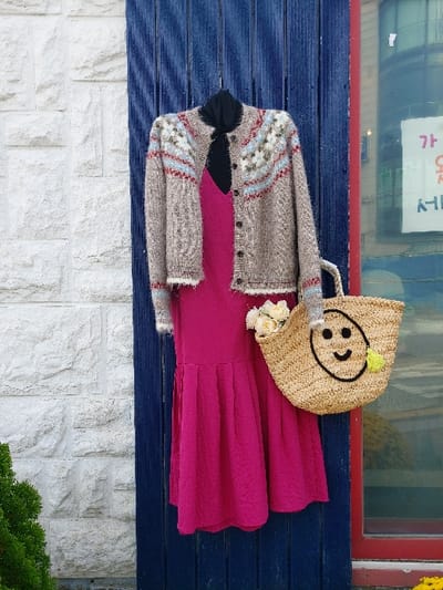 The image depicts an outdoor clothing and accessory display, presumed to be in 강화군, South Korea. A black high-neck top is layered under a fuchsia-colored sleeveless dress. Over these, a beige knitted cardigan with a Fair Isle-style pattern in light blue, red, and off-white is buttoned down the front. The cardigan features fuzzy white trim on its cuffs and hem. To the right, a light brown woven straw tote bag is suspended, adorned with a black embroidered smiley face and a small yellow pom-pom. This bag contains several cream-colored artificial flowers. The display is set against a dark blue vertical corrugated panel. To the left of the panel, a white textured stone wall is visible. To the right of the panel, a window with a red frame is present, reflecting a sign with Korean characters and displaying yellow elements at its base. A small portion of a green shrub is visible in the bottom left corner.