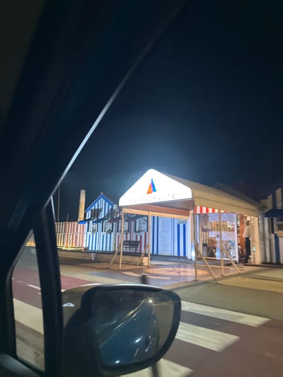 A nighttime view from inside a car, showing a side mirror reflecting a dark road with white and red stripes. Adjacent is a collection of small buildings adorned with blue and white stripes, resembling beach huts. A brightly lit, triangular sign is visible above, featuring colorful graphics. A larger building with a red and white striped awning stands slightly behind the huts, suggesting a commercial establishment.