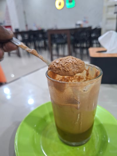 A close-up shot focuses on a glass of iced coffee placed on a green saucer. A spoon laden with frothy coffee sits above the glass, with a portion of a person's hand visible on the left edge of the frame, holding the spoon. The background suggests an indoor dining setting with tables and chairs, and in the far background, a blurry sign with yellow and green circles can be seen. The overall ambiance points to a cafe or restaurant in Keuniree, Indonesia.