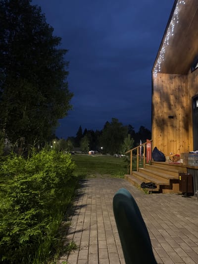 The image depicts an outdoor scene at dusk, likely in the vicinity of Doma 1096 Km, Russia. A wooden building with a covered porch and steps is visible on the right. String lights are draped along the edge of the building's roof. A paved pathway extends from the foreground, leading towards the building and a grassy area. A dense green hedge lines the left side of the pathway. In the background, there is a field with scattered trees and some distant structures, including what appears to be a small building or shelter. The sky is a deep blue, indicating the transition from day to night. There are no people visible in the image.