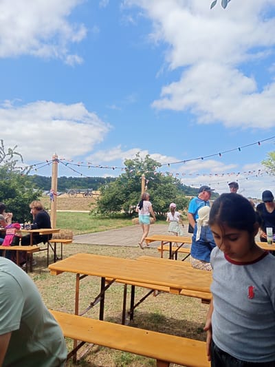 The image depicts an outdoor gathering in Azannes-et-Soumazannes, France, likely a local fair or festival, on a sunny day with scattered clouds. In the foreground, a young girl in a grey t-shirt with a red logo is looking down. To her left, a wooden picnic table and bench are visible, partially shaded. Further back, other people are seated at picnic tables and walking around. The scene includes trees, a wooden walkway, and a decorative string of colorful lights and small flags stretched between poles. In the background, a vintage wagon is parked on a dry, grassy area, with rolling hills covered in trees in the distance. The overall atmosphere is casual and festive.
