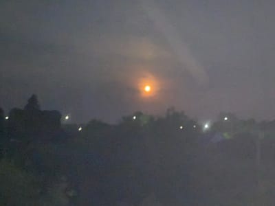 The image shows a hazy, dimly lit outdoor scene, likely taken at dusk or night. A bright, orange-yellow orb, possibly the moon or a setting sun, is visible in the upper center of the frame, partially obscured by clouds or atmospheric haze. The foreground is dominated by dark, silhouetted shapes that appear to be trees or foliage. Scattered lights, appearing as small white or yellow dots, are visible along the horizon, suggesting the presence of buildings or streetlights in the distance. The overall impression is one of a quiet, possibly rural or suburban setting, with limited visibility due to the atmospheric conditions. The image does not contain any recognizable landmarks or features that definitively place it in Polonne, Ukraine.
