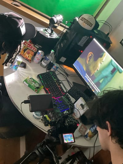A gaming setup sits on a round table. A computer monitor displays a face and French text. A black gaming keyboard with RGB lighting and a second keyboard are visible. A camera is set up on a tripod. Various cans, bottles, and snack packages like "Pizza Potato" are scattered around. A hat with "DEFOUGUE" is on the computer. The scene is framed by a green screen on the left. A person's hair obscures the upper right and another's head with dreadlocks is in the lower left.