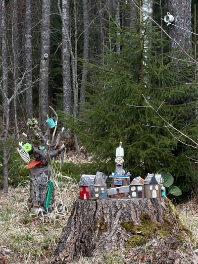The image shows a forested area, possibly in Glubokoe, Russia, based on the prompt. A tree stump dominates the foreground, adorned with a miniature village of small, wooden buildings. The houses are varied in design and color, creating a whimsical effect. To the left of the stump and attached to the base, found objects like miniature teacups and plastic toys are affixed to the tree. Behind the stump is a dense forest of thin trees with bare branches, and another evergreen tree. The environment is natural with grass and debris around the tree stump.
