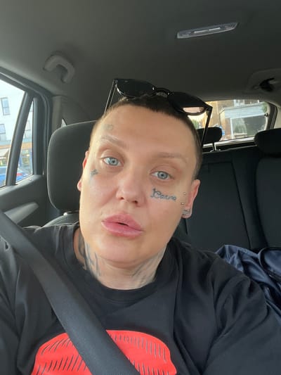 A pale-skinned individual with visible tattoos on their face and neck is sitting in what appears to be the driver's seat of a car. The individual is wearing a black t-shirt and a seatbelt. A pair of sunglasses is perched on the top of their head. The car's interior is visible, including the headrest and the rearview mirror area. Outside the car window, a building is visible, suggesting an urban or suburban setting. The specific location in Epping Forest, United Kingdom, cannot be determined from the image alone.