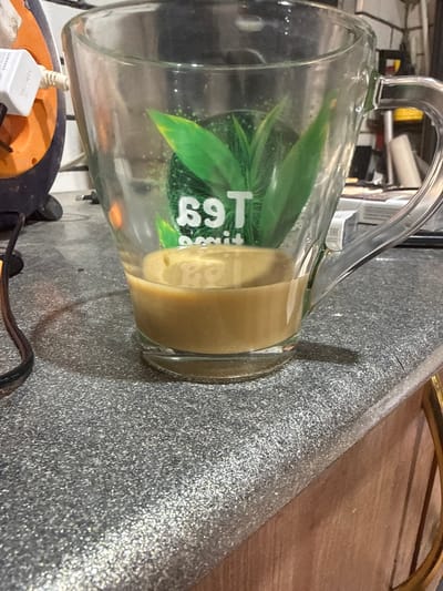 A close-up, eye-level shot captures a clear glass mug containing a light brown beverage, possibly coffee or tea, with some condensation on the interior. The mug sits on a speckled gray countertop. A part of a black cord is visible on the left side of the image, and an orange and black electronic device with a white "SAMSUNG" plug is situated behind the mug. The background is blurred, but hints of shelving and various objects can be discerned. The lighting suggests an indoor setting, likely a kitchen or workspace. There is no indication of the city of Bryansk, Russia, or any specific geographical location in the image.