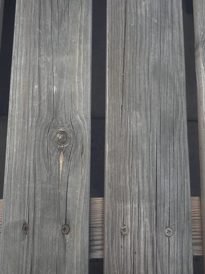 The image shows a close-up, overhead view of weathered wooden planks, likely part of an outdoor structure such as a deck or fence. The wood is a muted gray color with visible grain patterns, cracks, and knots. Two of the planks have rusted screws embedded in them, one on the left and one on the right, both near the bottom of the frame. The planks are arranged vertically and are separated by dark shadows, suggesting they are either spaced apart or are positioned above a darker surface. The overall impression is one of aged, rustic wood. The location in the city of San Lorenzo de Tormes, Spain, is not directly evident from the image itself, as the scene is a generic depiction of weathered wood.