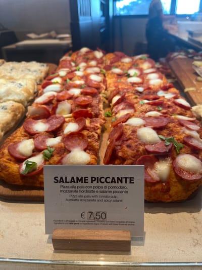 The image displays a close-up of two long, rectangular pizzas, known as "pizza alla pala," presented on wooden serving boards, likely in a food establishment in Dorno, Italy. The pizzas are generously topped with a red tomato pulp base, numerous round slices of spicy salami, and visible dollops of melted fiordilatte mozzarella cheese. Small green herb leaves, possibly basil or oregano, are scattered across the toppings.
To the right, a white price tag or description card, held by a wooden base, provides details about the item. The prominent heading reads "SALAME PICCANTE" (Spicy Salami). Below, the description states in Italian: "Pizza alla pala con polpa di pomodoro, mozzarella fiordilatte e salame piccante," followed by an English translation: "Pizza alla pala with tomato pulp, fiordilatte mozzarella and spicy salami." The price for this item is clearly marked as "€ 7,50." Smaller text at the bottom provides information about ingredients and allergens, noting that the product's base was frozen at source. The scene is brightly lit, suggesting an indoor setting during the day, showcasing the appetizing appearance of the pizzas.