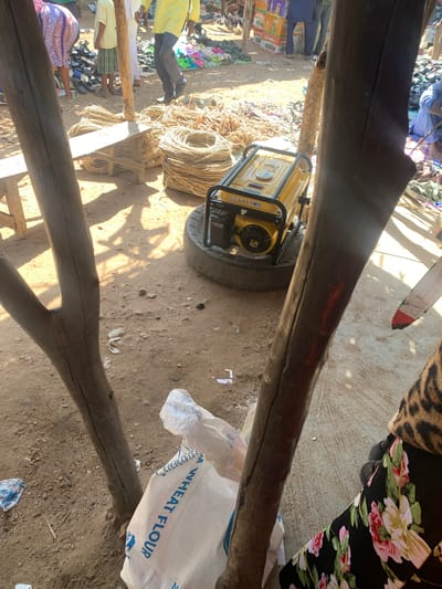 This image captures a bustling marketplace scene, likely in Dull A, Nigeria. The primary subjects are several people, though their faces are mostly obscured or out of focus. They are dressed in everyday clothing, suggesting a typical market day.

Various objects are scattered around the scene, indicating commercial activity. A prominent yellow generator is visible, suggesting it's being used to power something or is for sale. Large coils of rope are piled up, along with what appear to be bags of goods, one of which has the text "WHEAT FLOUR" clearly visible in blue lettering. In the background, there are stacks of colorful items, possibly textiles or other merchandise. The ground is dirt, typical of an outdoor market.

The scene is lit by bright sunlight, casting distinct shadows, suggesting it is daytime, likely mid-morning or afternoon. The overall atmosphere appears to be one of activity and commerce. The presence of both goods for sale and a generator implies that this is a working market where essential services might be supported by such equipment. The wooden beams in the foreground frame the scene, suggesting a covered or semi-covered stall area. No specific emotions are discernible from the image.