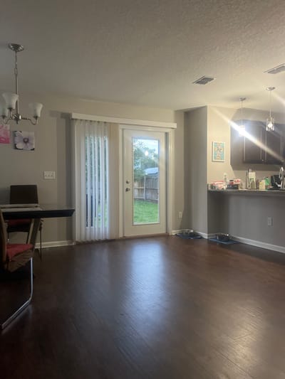This is a full-shot, eye-level, indoor photograph of a dining and kitchen area in Jacksonville, United States of America. The room has dark wood flooring and light grey walls. In the foreground, a dark dining table with chrome legs and a dark chair with a red cushion are visible on the left. Two floral art pieces hang on the wall behind the table. A chandelier with three lights hangs from the ceiling above the dining area.

A sliding glass door with vertical blinds leads to a backyard with a green lawn and a wooden fence. Two ceiling vents are visible in the ceiling. In the background, a kitchen counter with dark cabinets above and to the right is present. Several items are on the counter, including bottles, containers, and a coffee maker. Two dog bowls on blue mats are placed on the floor near the kitchen counter. The lighting appears to be artificial, with overhead lights and light coming from the backyard.