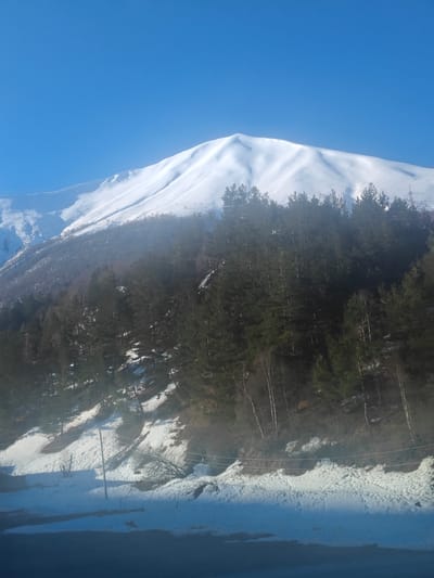 The image displays a vibrant winter mountainous landscape, likely in Almasiani, Georgia, under a clear, bright blue sky. A large, snow-covered mountain with gently contoured slopes dominates the background, bathed in sunlight. Below the prominent peak, a dense forest of dark green evergreen trees covers the mid-ground, extending down the mountain's side. A slight haze or mist partially obscures the trees in the middle distance.
In the foreground, the ground is covered in patches of snow, with clear footprints visible, suggesting recent human or animal activity. A dark, seemingly wet or paved surface, possibly a road or path, runs along the very bottom of the frame. A wooden utility pole with overhead lines stands on the snow-covered slope on the right side, indicating some infrastructure in the natural setting. The time of day appears to be late morning or early afternoon, characterized by strong sunlight and clear visibility. The weather is crisp and cold, typical of a sunny winter day in a snowy region. No discernible text is visible in the media file.
